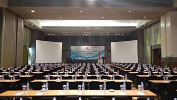 Un salón de conferencias con filas de mesas, cada una con botellas de agua y vasos, y una pantalla grande al frente.