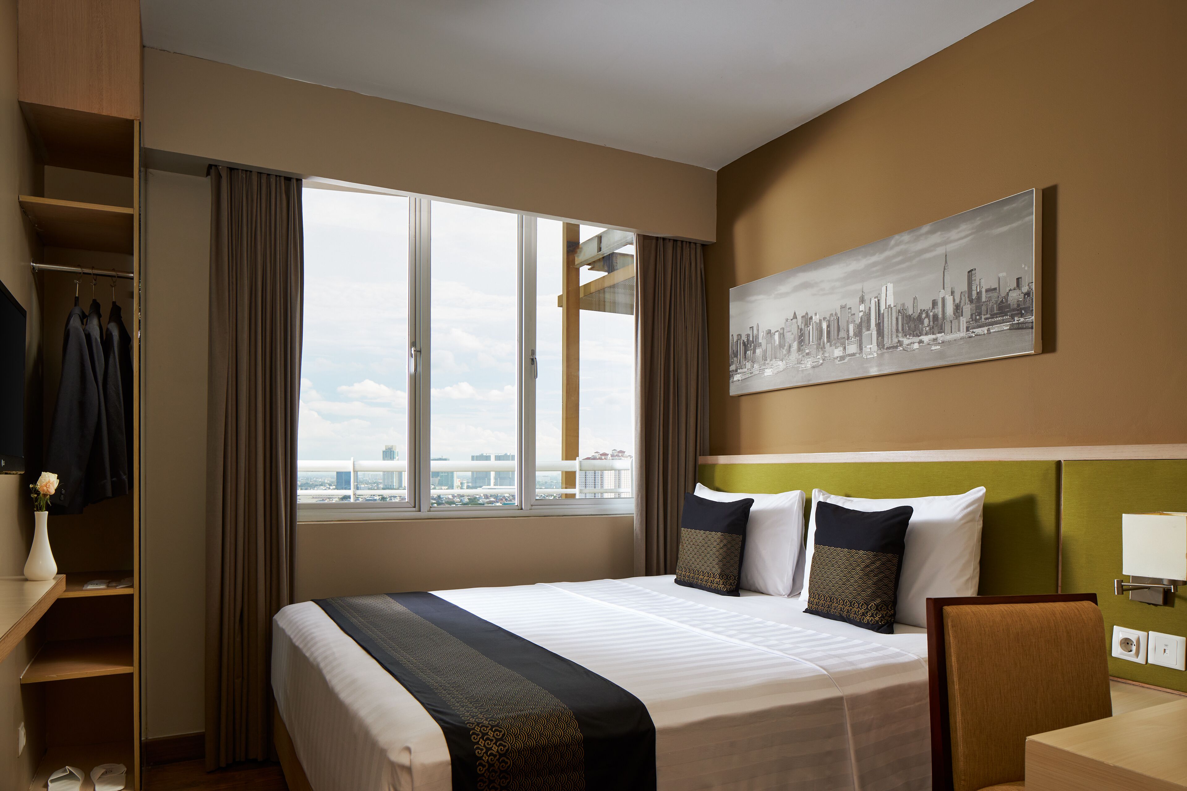 A hotel room with a large bed, a cityscape painting, a wardrobe, and a view of the city through the window.