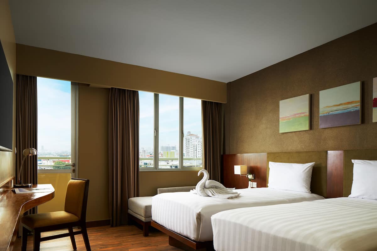 Ein Hotelzimmer mit zwei Betten, einem Schreibtisch, einem Sessel und einem großen Fenster mit Blick auf die Stadt.