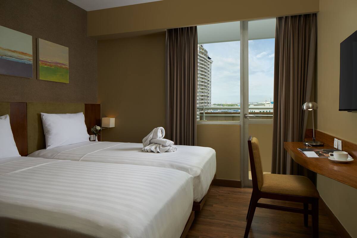 Ein Hotelzimmer mit zwei Betten, einem Schreibtisch mit Stuhl, einem Fernseher und Blick auf eine Stadtlandschaft durch große Fenster.