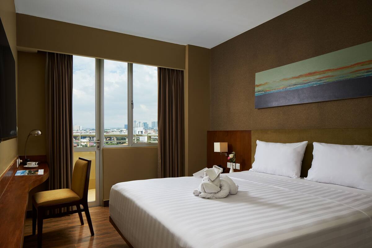 Ein Hotelzimmer mit einem großen Bett, einem Schreibtisch, einem Sessel, einem Fenster mit Vorhängen und einem Bild an der Wand.