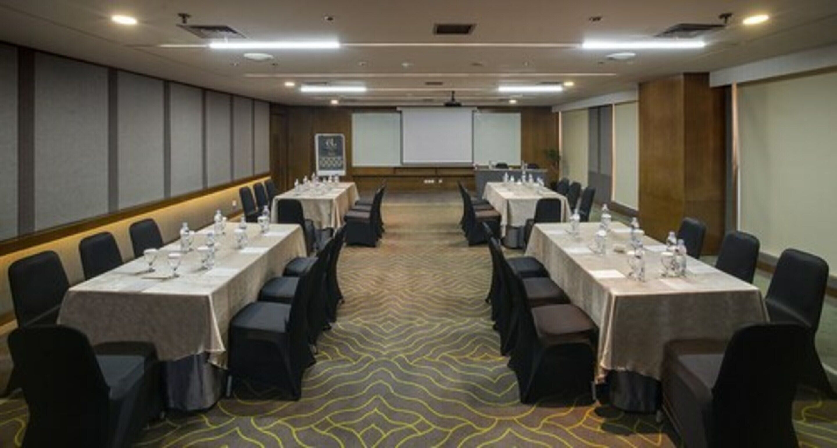 Un salón de conferencias con dos largas mesas, cada una rodeada de sillas, equipado con botellas de agua y vasos.