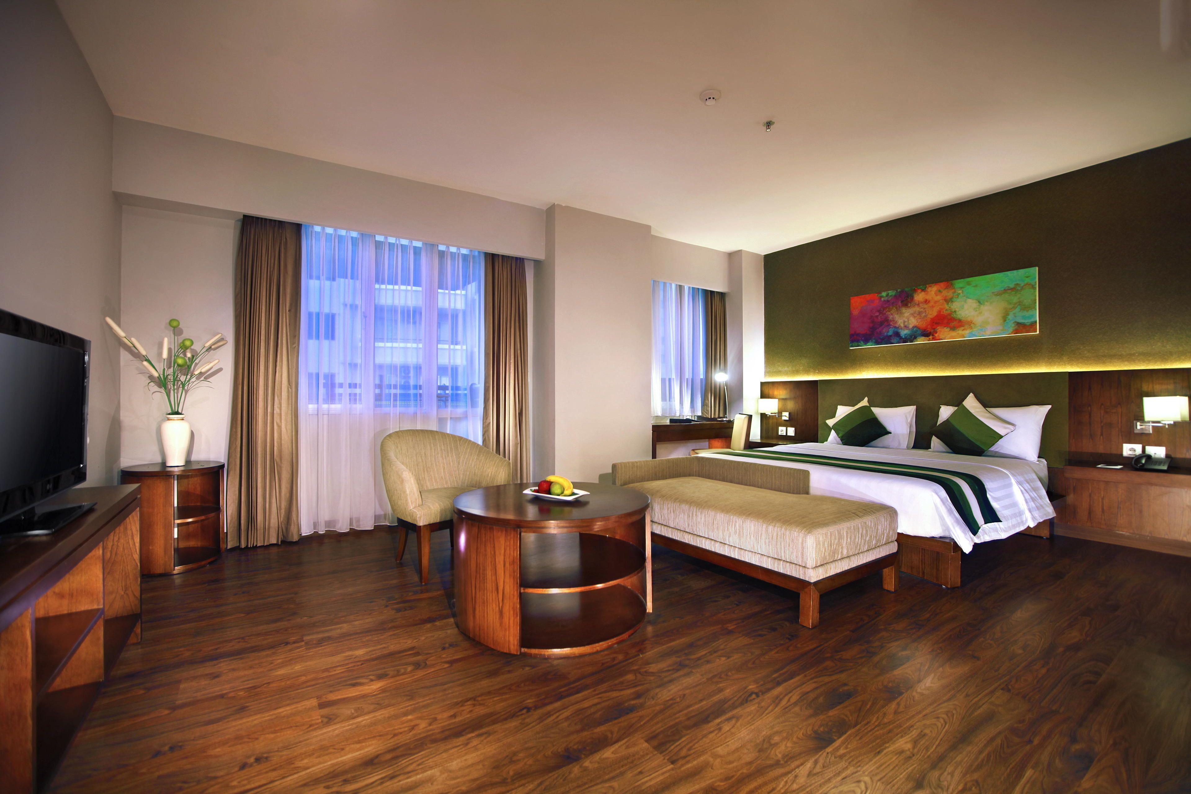 Hotelkamer met een bed, een bank, een televisie, een ronde houten tafel en een groot raam met gordijnen.