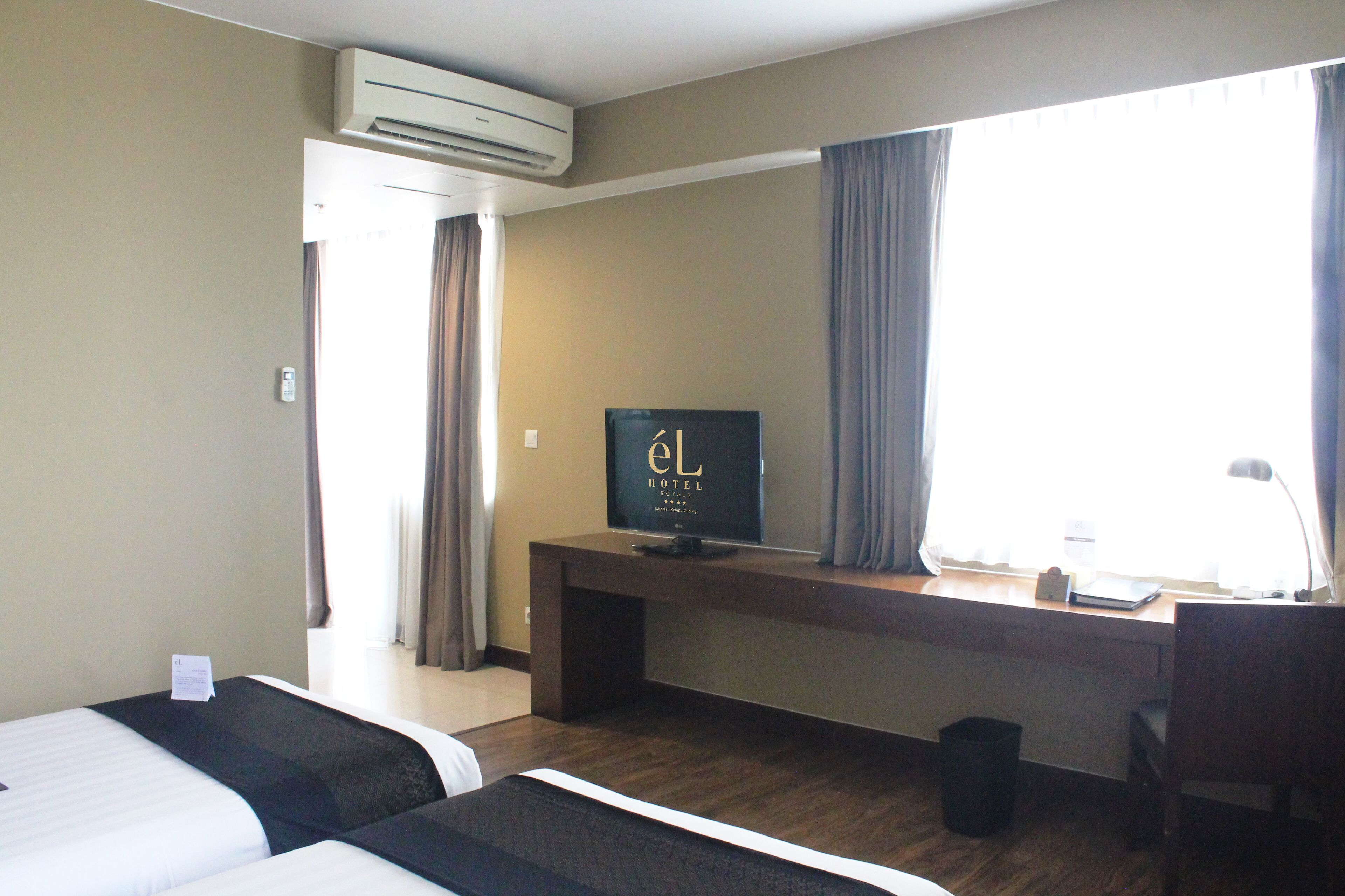 Een hotelkamer met twee bedden, een bureau met een televisie, een raam met gordijnen en een airconditioner.