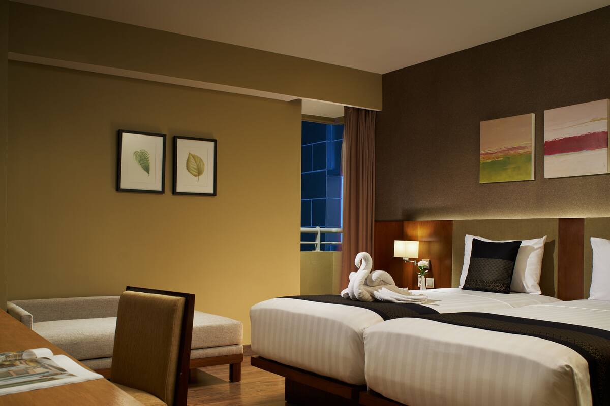 Ein Hotelzimmer mit zwei Betten, einem Sofa, einem Sessel, gerahmten Bildern an der Wand und einem Fenster mit Vorhängen.