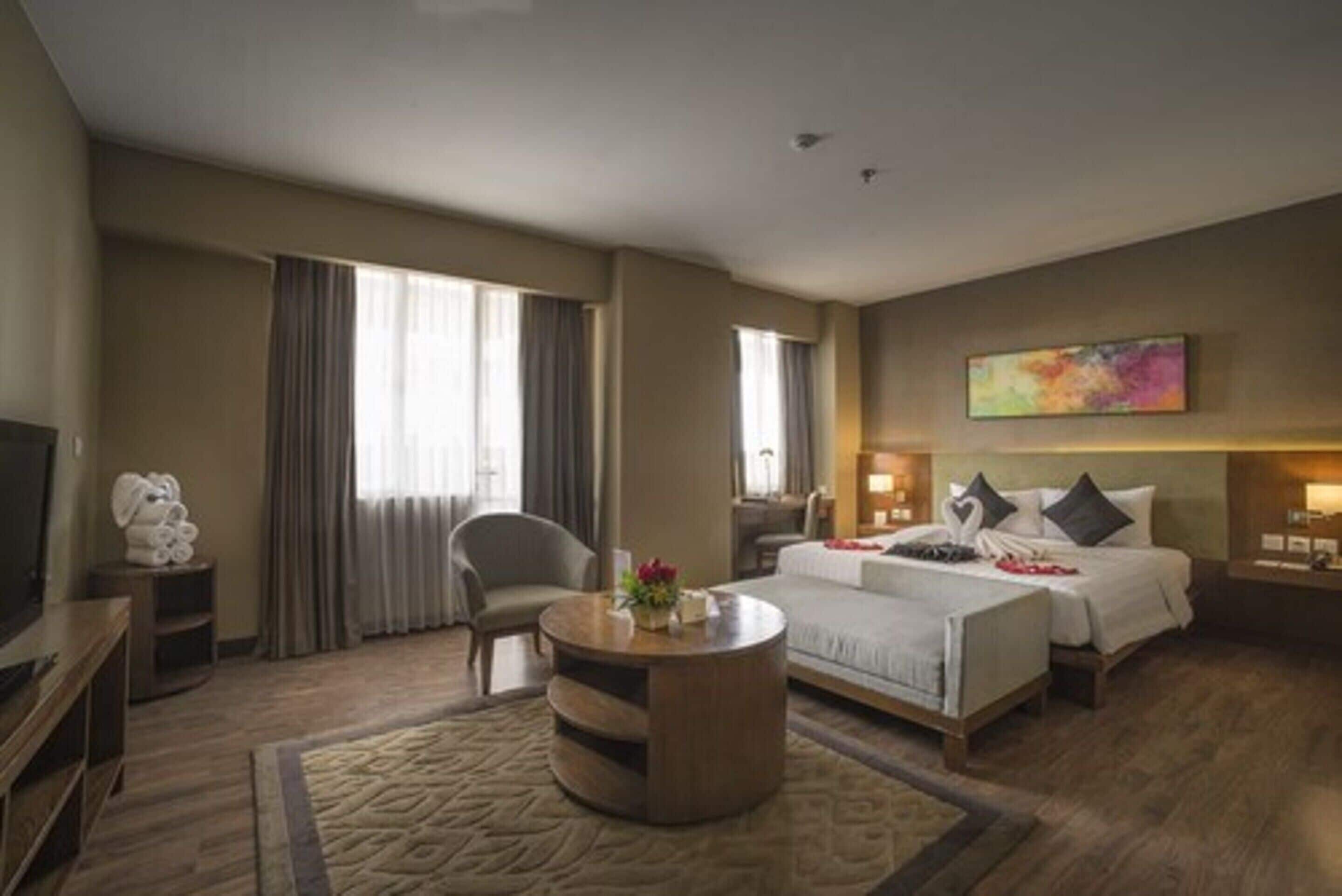 A hotel room with a bed, a round coffee table, a chair, a TV, and a painting on the wall.
