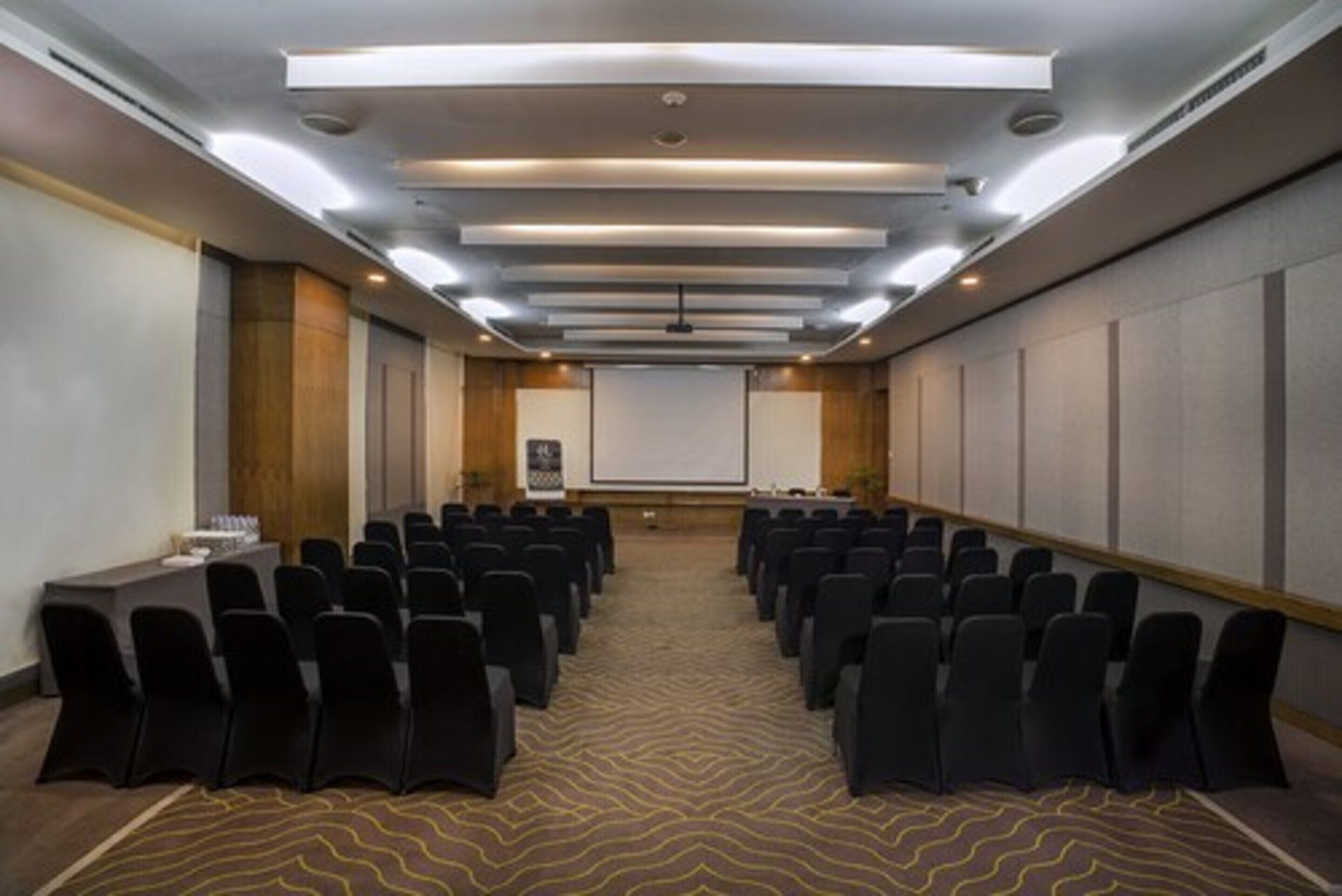 Una sala conferenze con file di sedie, uno schermo per proiezioni e un tavolo per presentazioni.