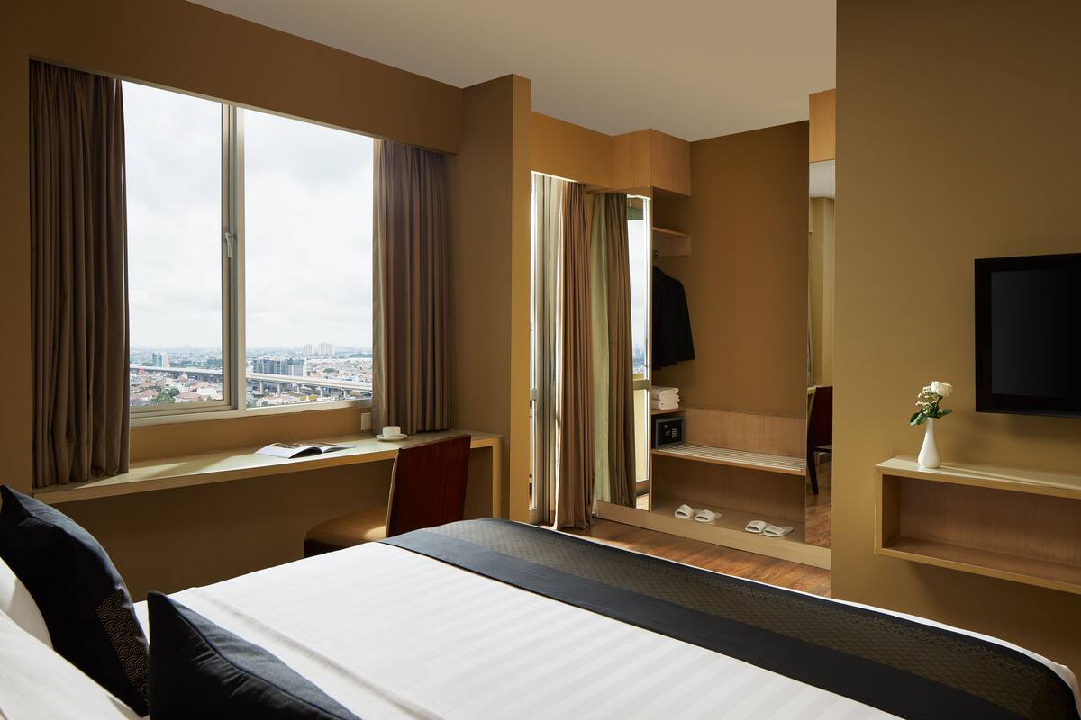 Ein Hotelzimmer mit einem großen Bett, einem Schreibtisch, einem Stuhl, einem Fernseher, einem Spiegel und einem Fenster mit Vorhängen.