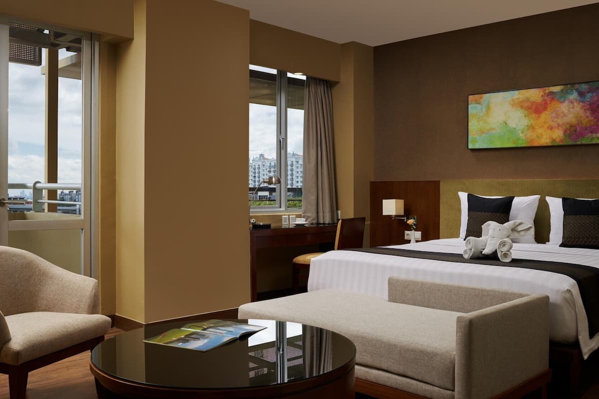 Ein Hotelzimmer mit Bett, zwei Sesseln, einem kleinen Tisch, einem Gemälde und einem Fenster mit Blick auf die Stadt.