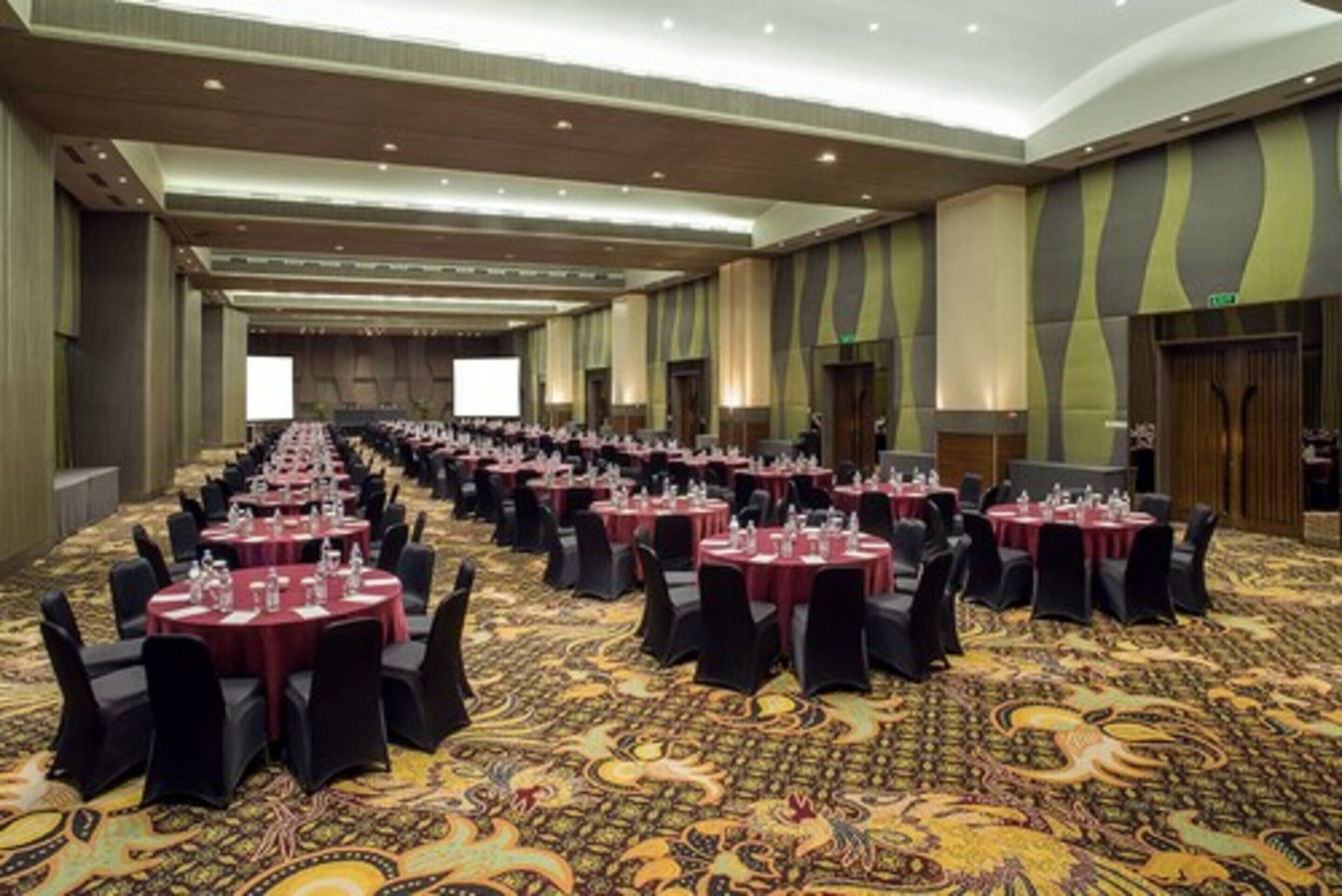 Un amplio salón de conferencias con filas de mesas y sillas, suelo alfombrado y techo alto.