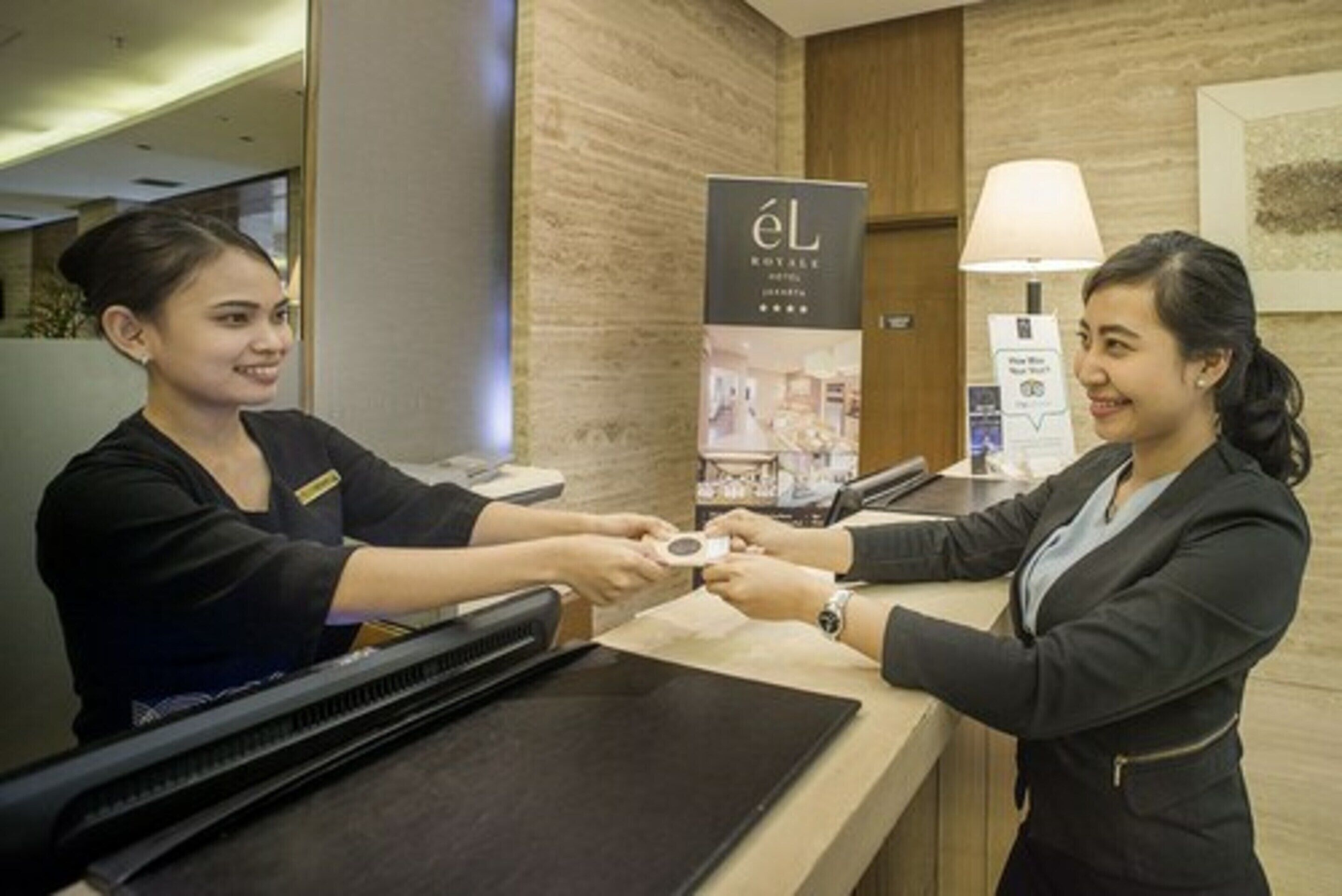 A hotel reception area with a reception desk, a digital screen, and a sign for 'eL Royal Hotel'.