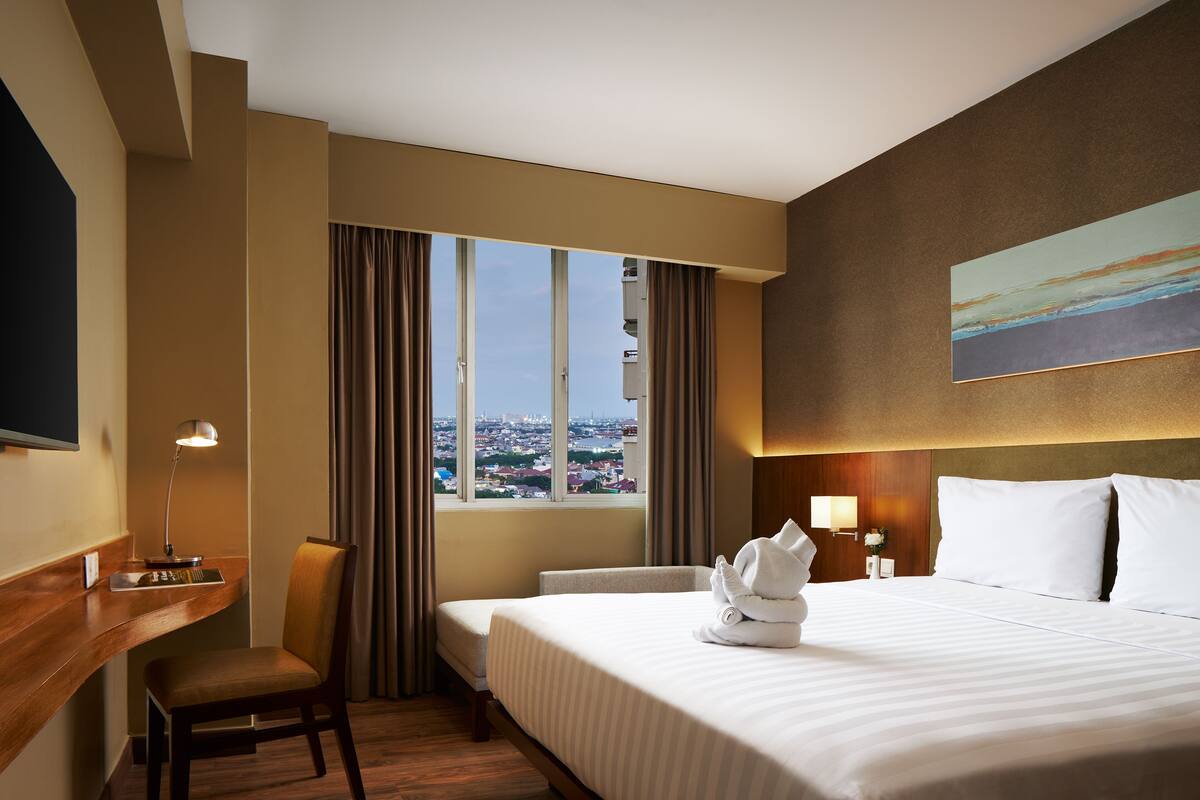 Ein Hotelzimmer mit einem großen Bett, einem Schreibtisch, einem Sessel, einem Fenster mit Vorhängen und einem Bild an der Wand.