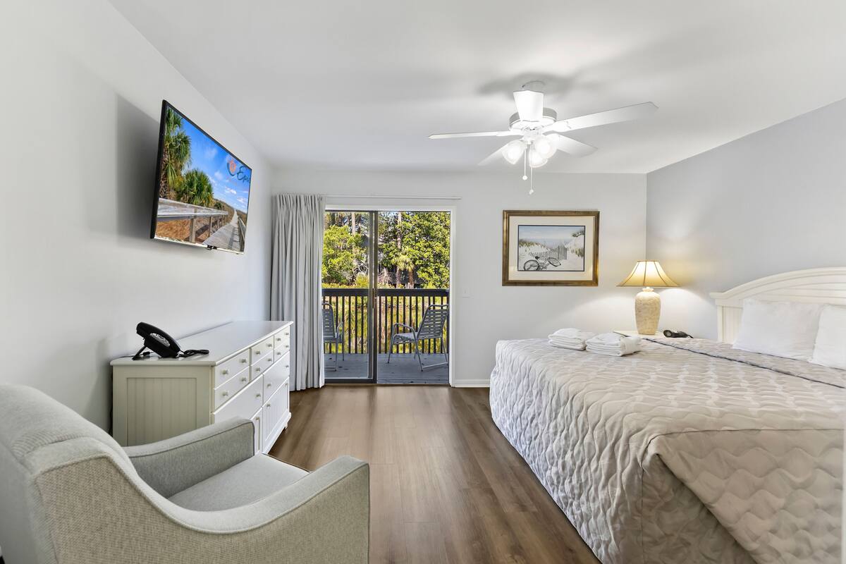A hotel room with a bed, a desk with a phone, a chair, a ceiling fan, a TV, and a balcony with a view of trees and a fence.