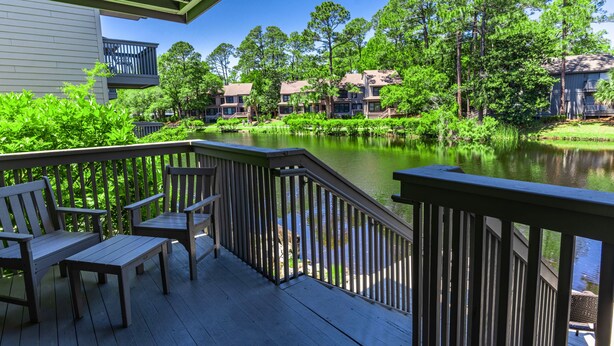 A wooden deck with benches overlooking a calm lake, surrounded by lush greenery and residential buildings. - Ocean Cove At Palmetto Dunes (Hilton Head Island)