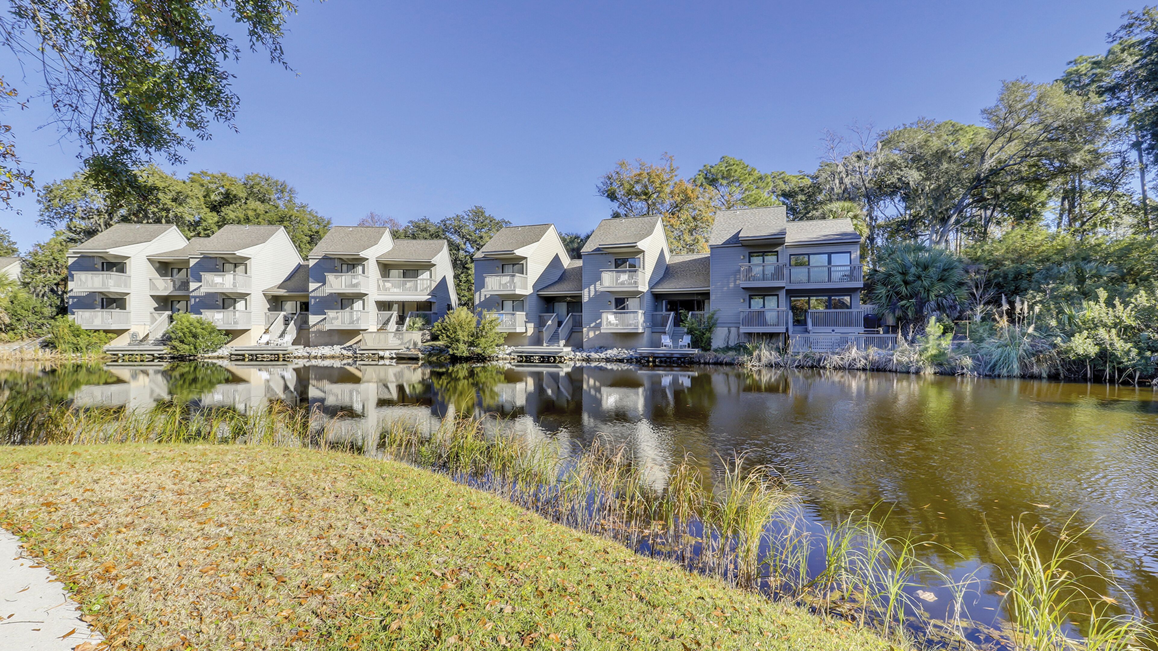 A row of multi-story residential buildings with balconies, a calm pond in the foreground, and surrounding greenery.