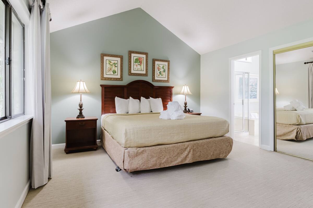 A bedroom with a bed, two bedside tables with lamps, a large mirror, and three framed botanical prints on the wall.