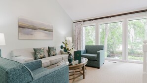 Una sala de estar con sofá, sillón y mesa de centro, ventanas amplias con vistas a la vegetación y un cuadro en la pared.