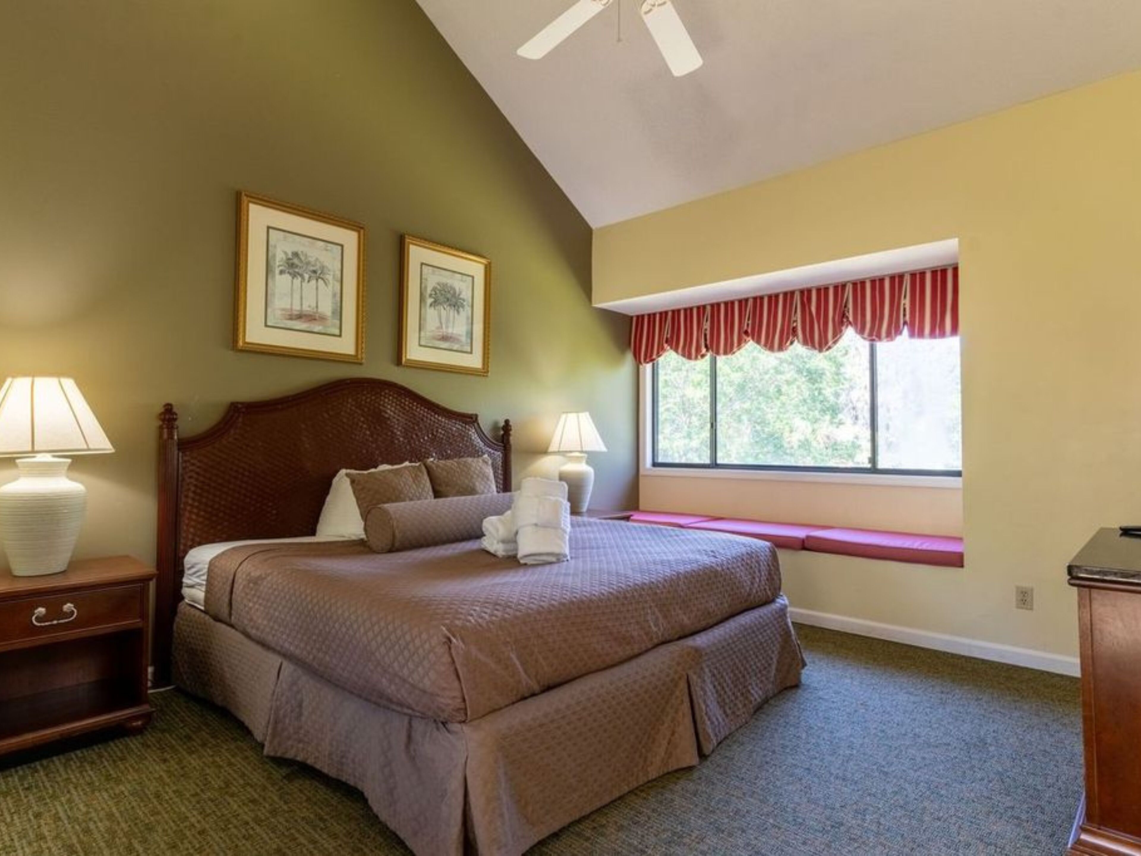 Une chambre à coucher avec un grand lit, deux lampes de chevet, une tête de lit en bois, une fenêtre agrémentée d’une lambrequin rouge et deux tableaux encadrés accrochés au mur.