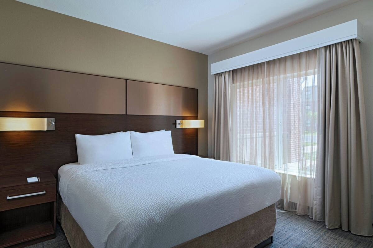 Habitación de hotel con una cama grande, cabecera de madera, mesita de noche y una ventana con cortinas.