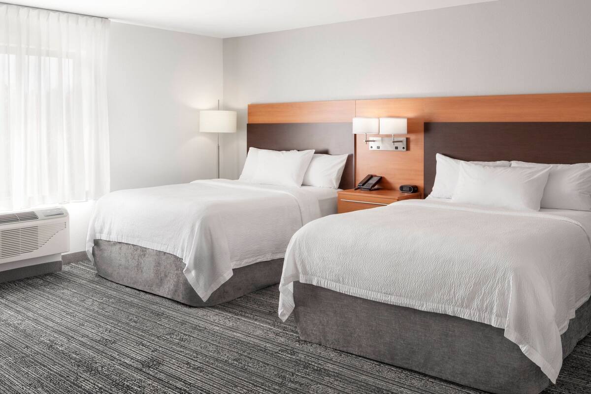 A hotel room with two beds, a bedside table with a lamp, a phone, and a wall-mounted light.
