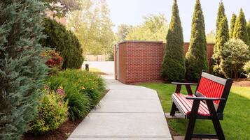 A pathway leading to a brick structure, surrounded by greenery and a bench with red cushions.
