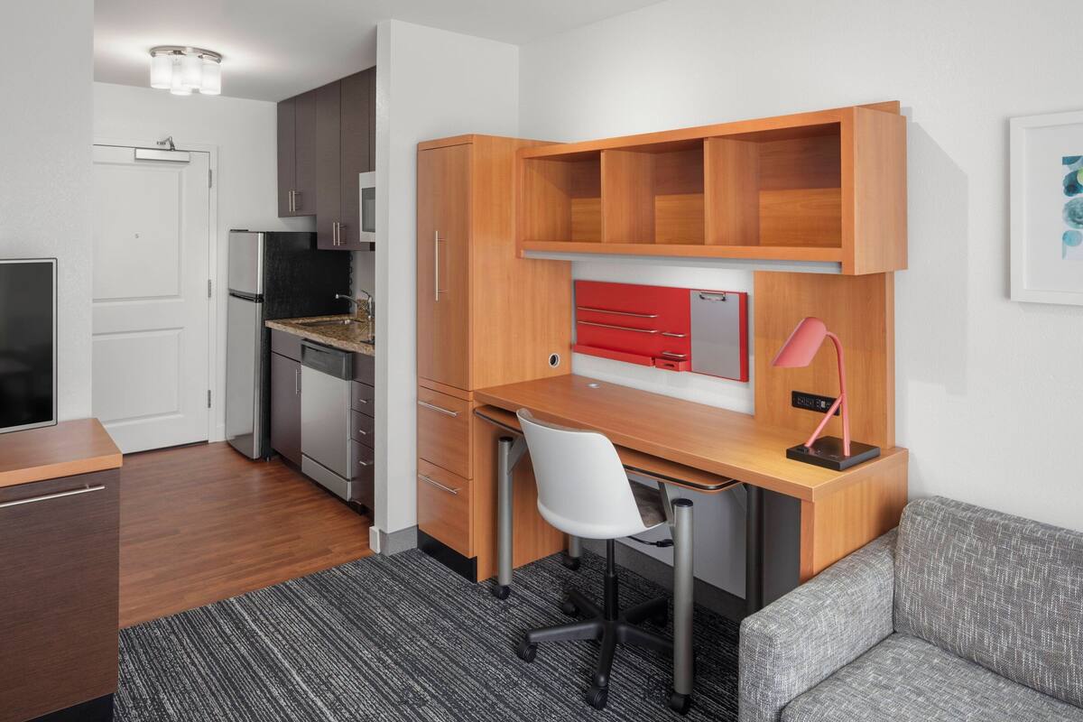 A compact kitchen with a built-in refrigerator, a wooden desk with a chair, and a grey sofa.