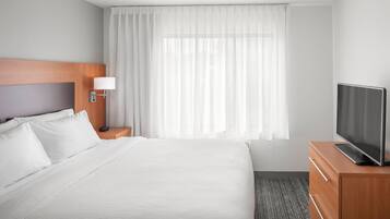 A hotel room with a large bed, a TV on a wooden dresser, a nightstand with a lamp, and a window with sheer curtains.