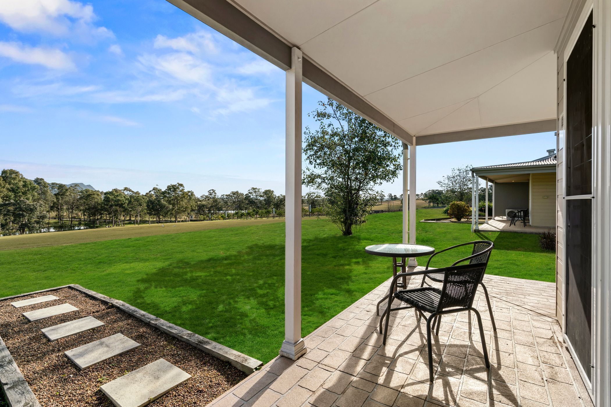 A covered patio with a table and chairs, a view of a well-maintained lawn and distant trees, and a building with a tiled roof.