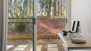 A room with a sliding glass door leading to a garden with trees and mulch. There is a table with plates, glasses, and a vase. - H Boutique Hotel (Pokolbin)
