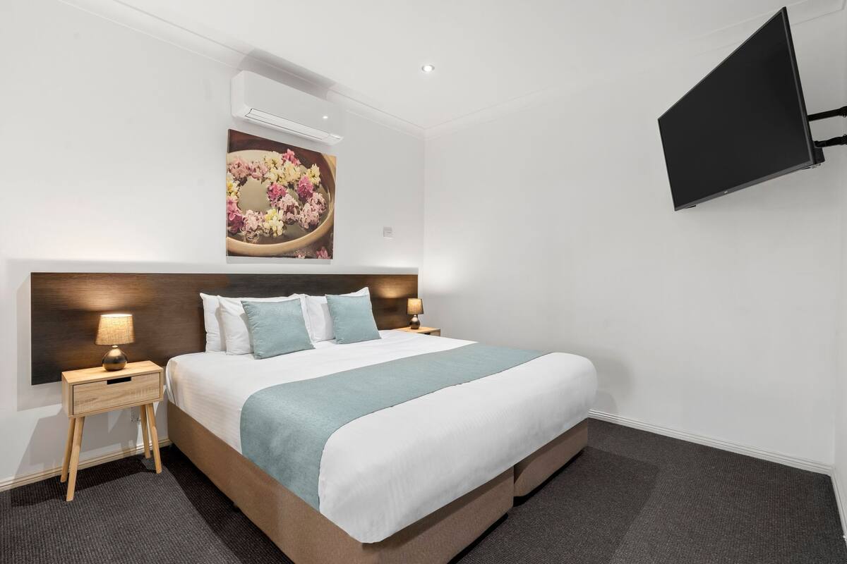 Ein Hotelzimmer mit einem großen Bett, einem flachbildfernseher an der Wand, einem kleinen Nachttisch mit einer Lampe und einem Gemälde über dem Bett.