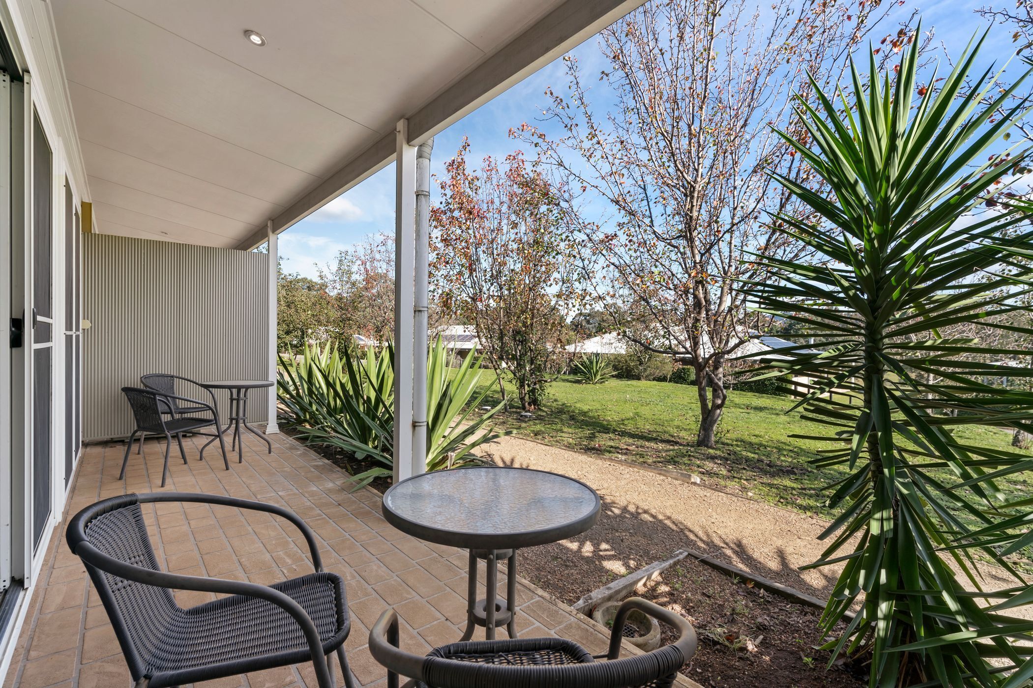 Un patio cubierto con mobiliario de mimbre, una mesita y vistas a un jardín con árboles y césped.
