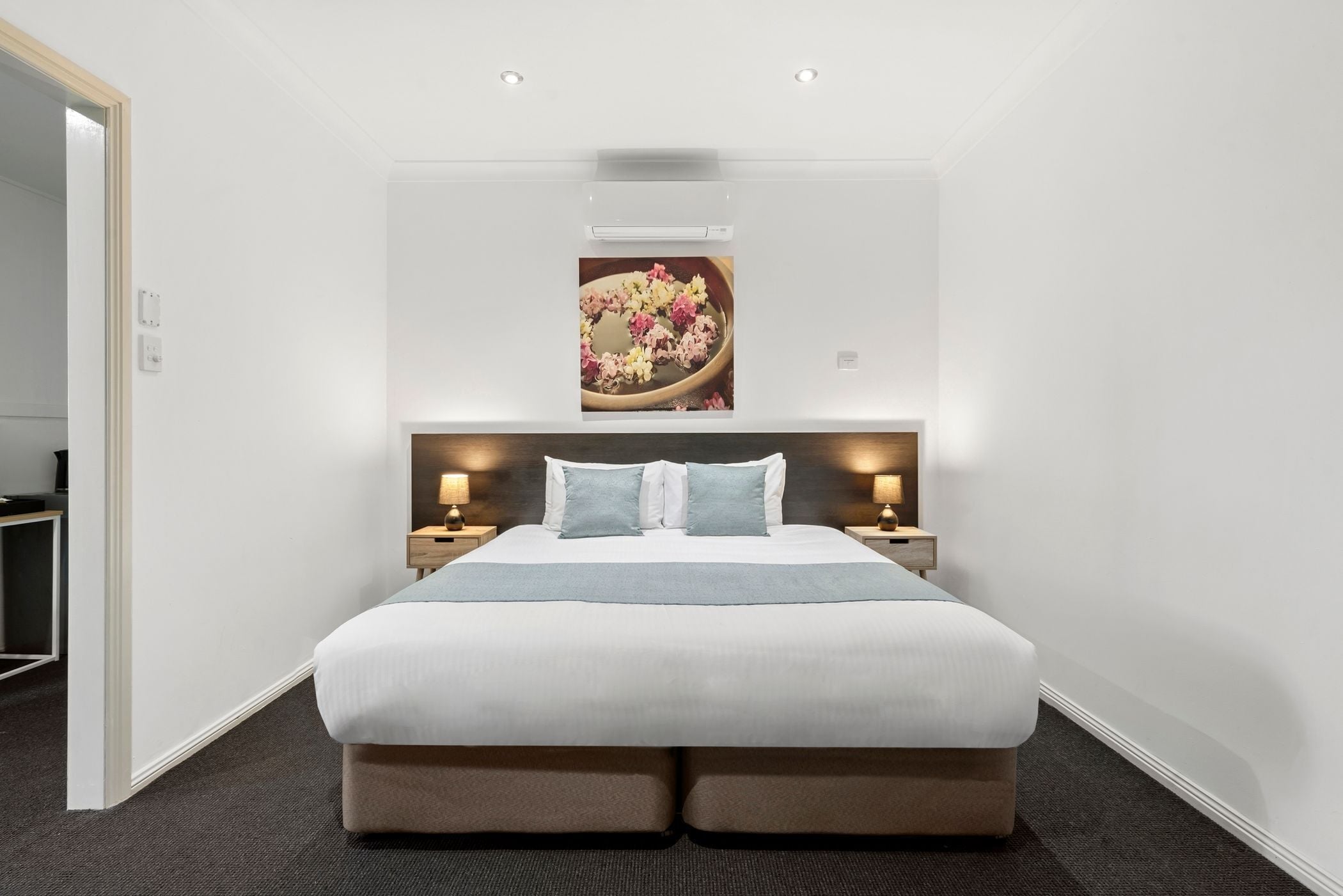 Une chambre d’hôtel moderne avec un grand lit, deux tables de chevet avec des lampes, un climatiseur fixé au mur et un tableau encadré au-dessus du lit.