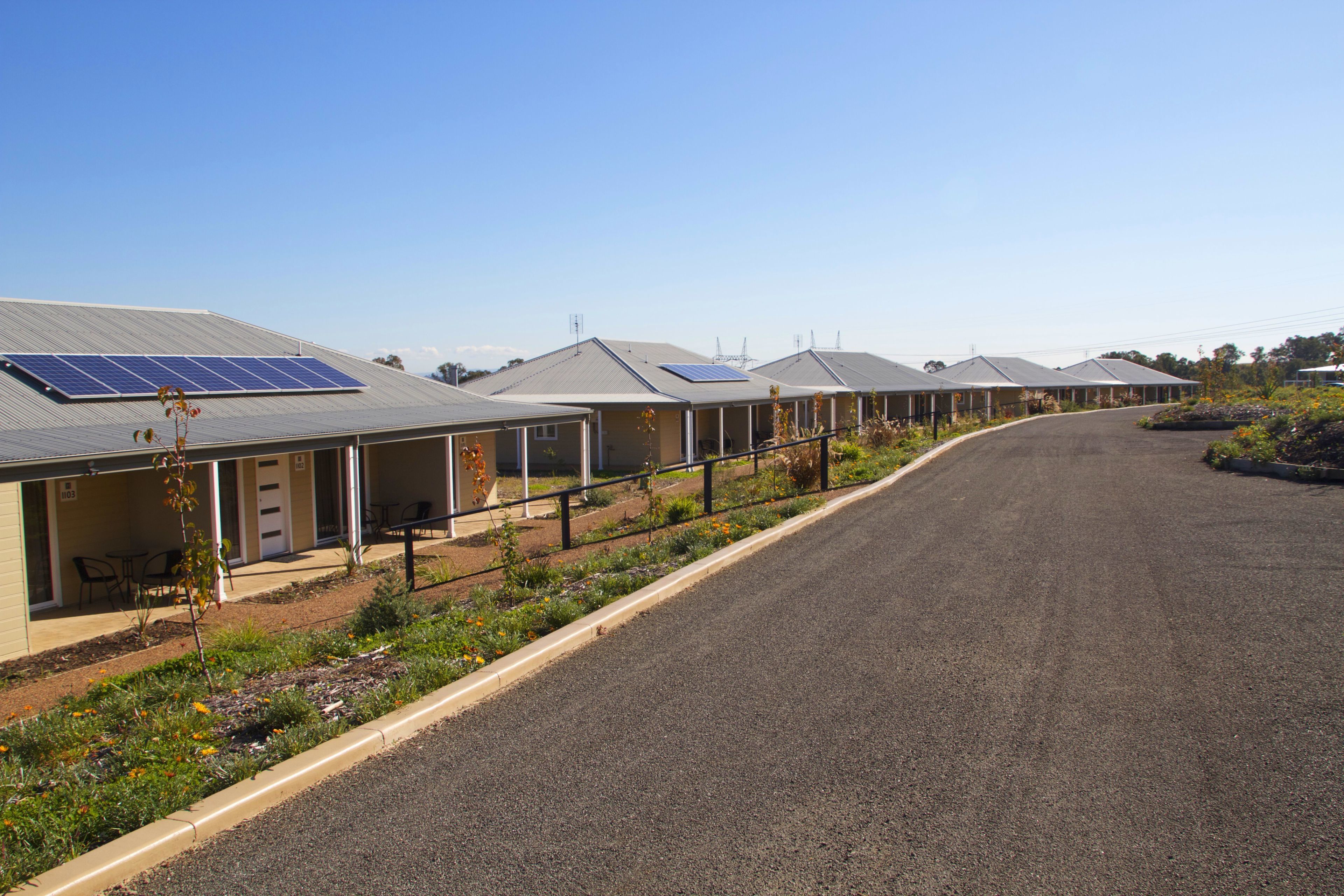 A row of single-story buildings with solar panels on the roofs, a paved road, and a landscaped area with flowers.