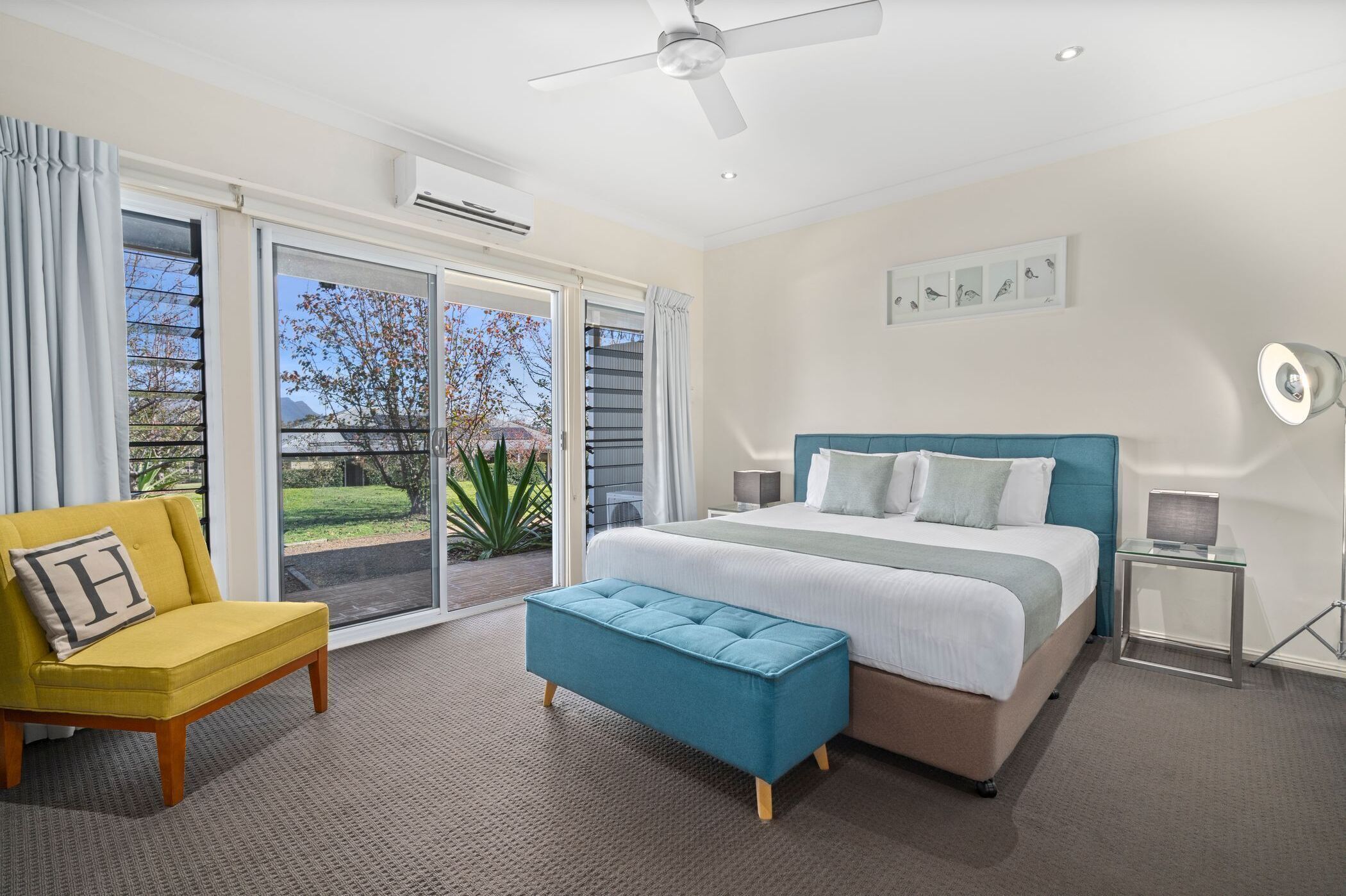A modern bedroom with a large bed, a yellow armchair, a blue ottoman, and a view of the outdoors through a sliding glass door.