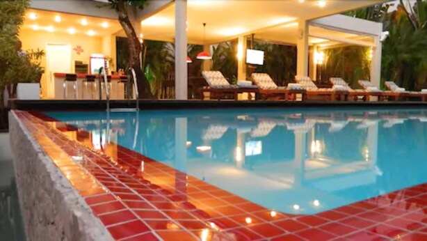 Une piscine dont les bords sont recouverts de carreaux, avec des chaises longues et un espace de détente couvert équipé de parasols.