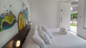 Une chambre d’hôtel avec un lit, des oreillers blancs, un mur orné d’œuvres d’art colorées, une porte portant le numéro 16 et une vue sur la verdure à l’extérieur.