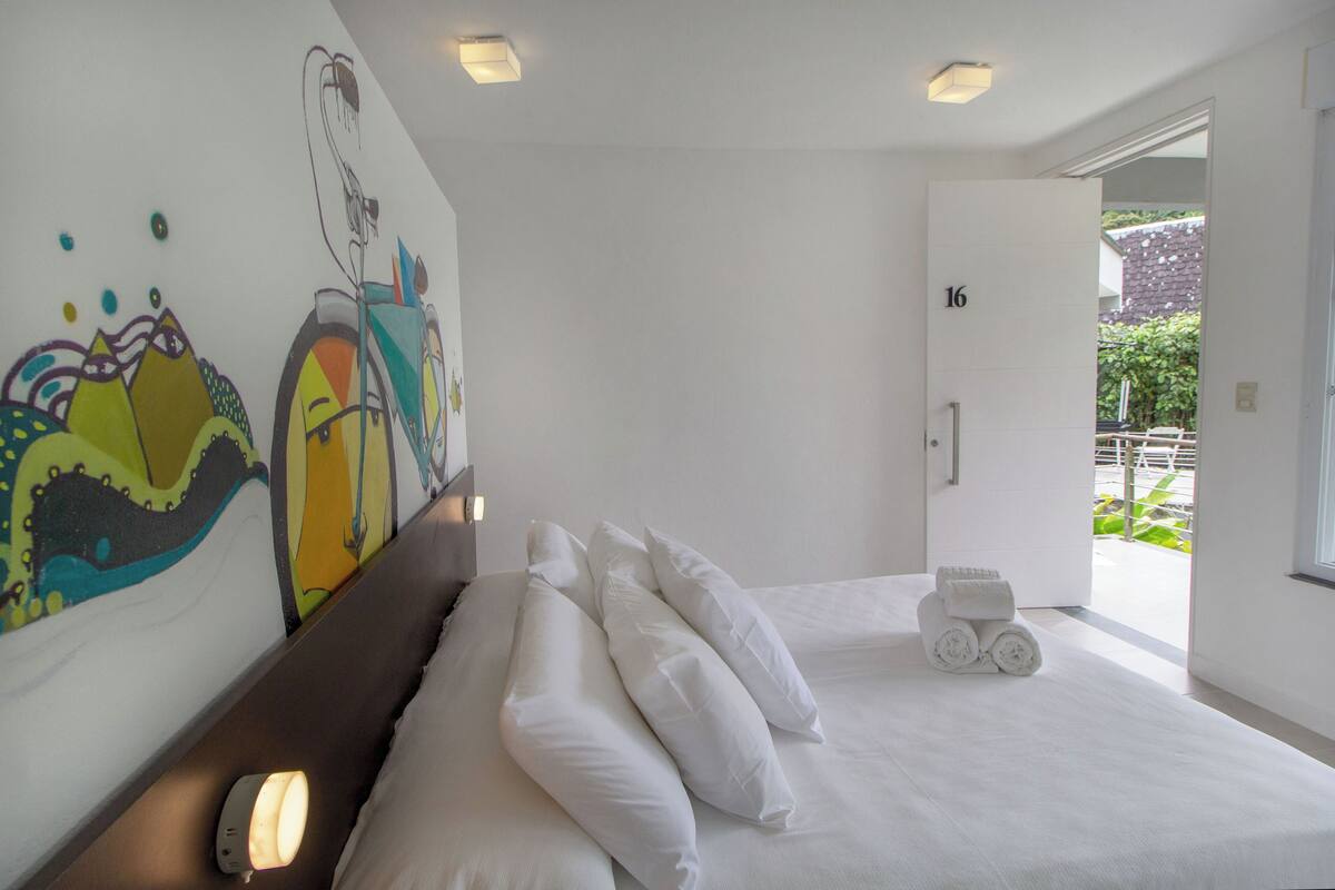 Quarto de hotel com cama, almofadas brancas, parede com arte colorida, porta com o número 16 e vista para áreas verdes lá fora.
