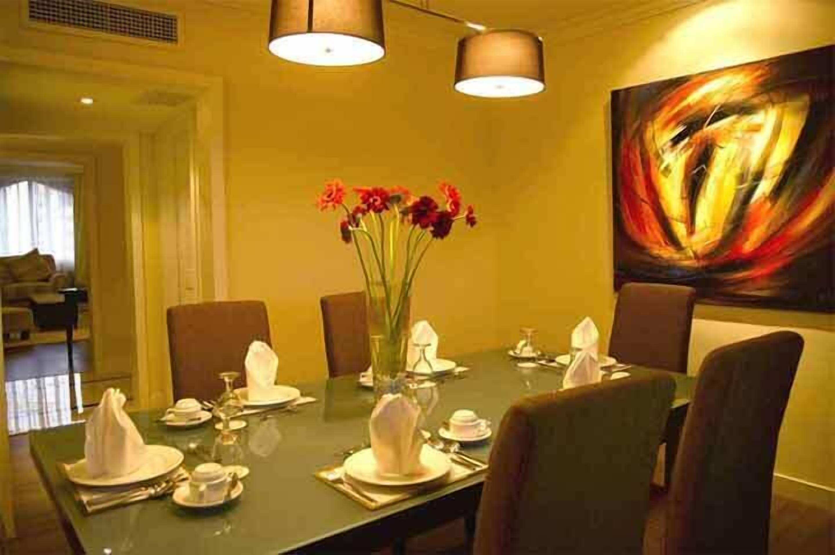 Zona pranzo con tavolo apparecchiato per quattro persone, un vaso di fiori rossi e un grande quadro astratto appeso al muro.