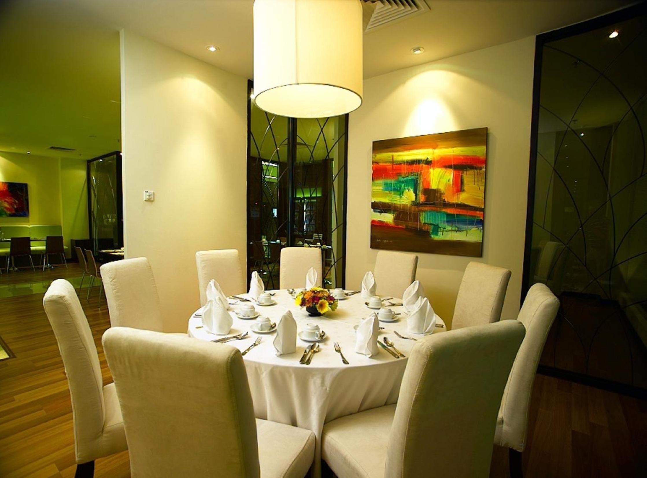 A dining area with a round table set for six, surrounded by chairs, with a large abstract painting on the wall and a window with a view of the cityscape.