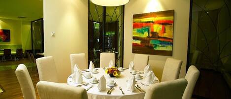 A dining area with a round table set for six, surrounded by chairs, with a large abstract painting on the wall and a window with a view of the cityscape.