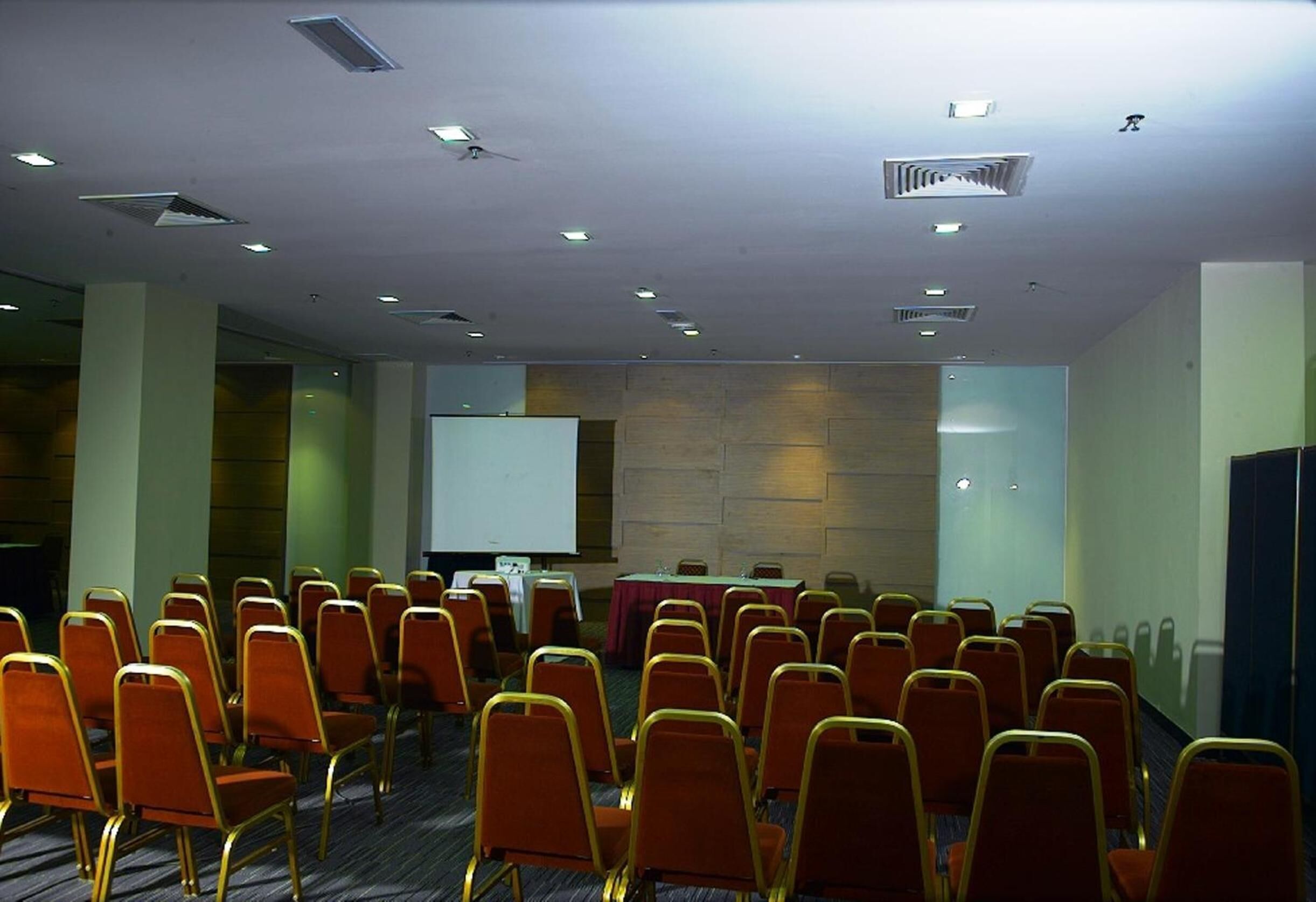 Une salle de conférence avec des rangées de chaises, un écran de projection et une table recouverte d’une nappe.