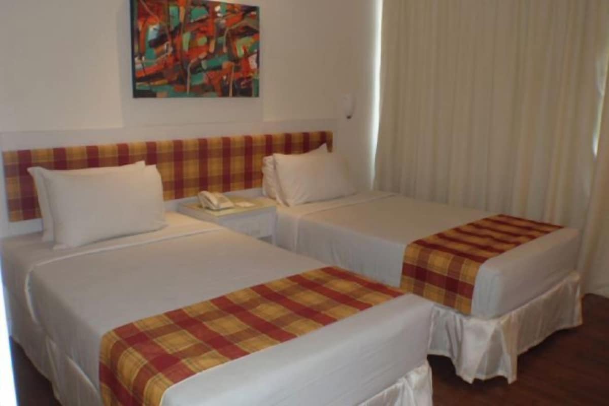 A hotel room with two beds, each with a white bedspread and a plaid blanket. There is a painting on the wall above the beds and a window with curtains.