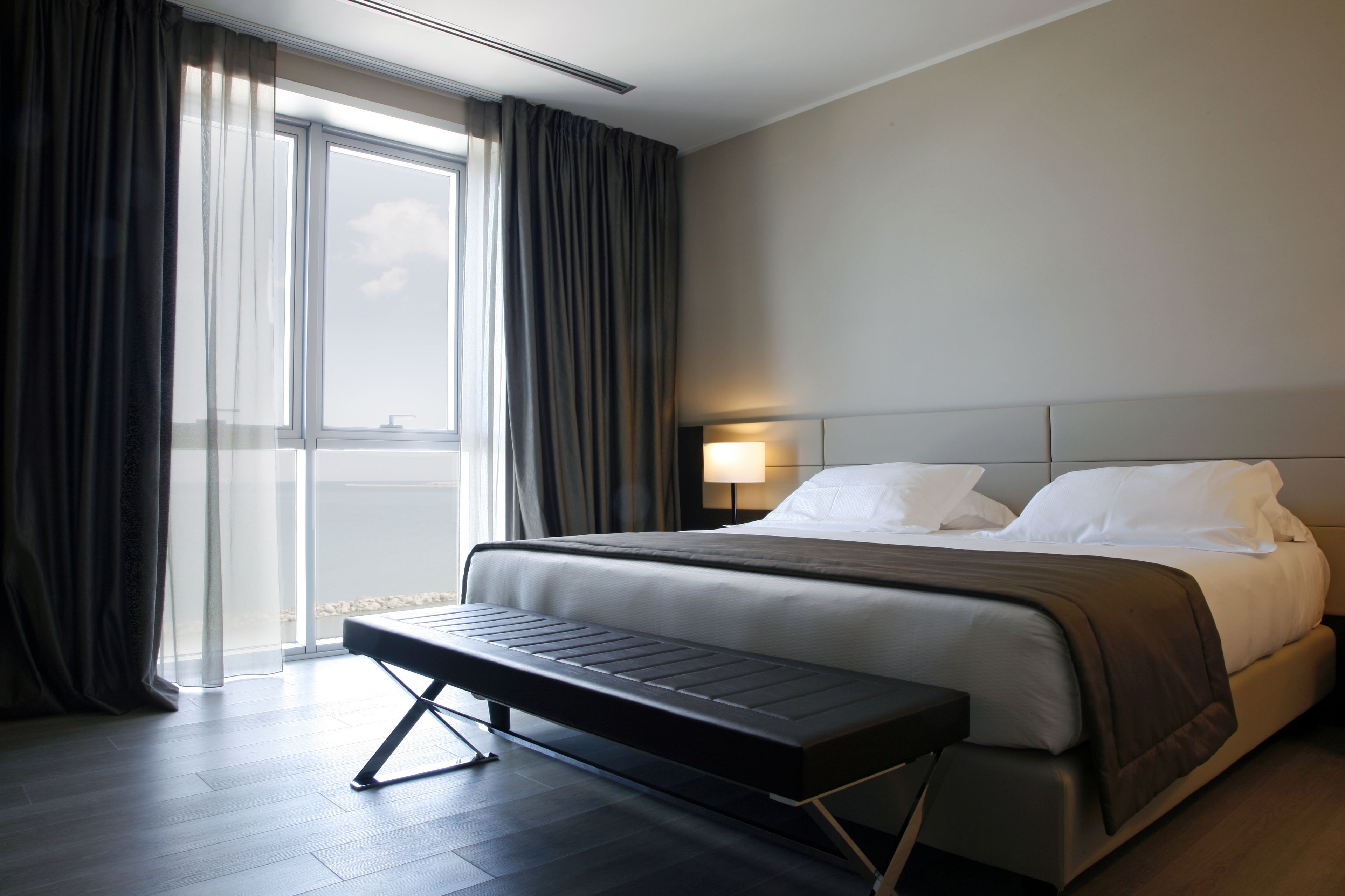 Ein modernes Hotelzimmer mit einem großen Bett, einer Bank, einem Nachttisch und einem Fenster mit Vorhängen.