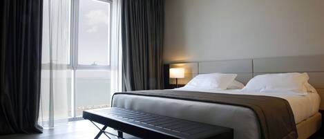 Ein modernes Hotelzimmer mit einem großen Bett, einer Bank, einem Nachttisch und einem Fenster mit Vorhängen.