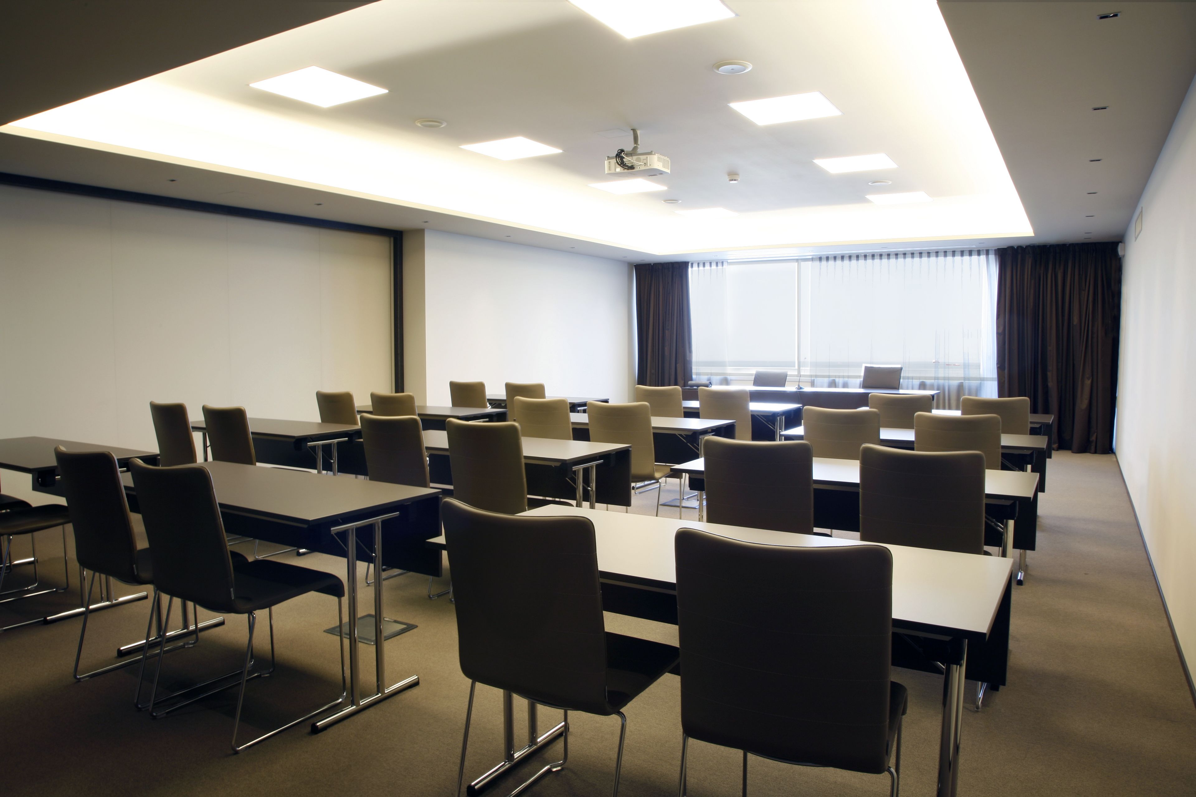 Une salle de conférence équipée de tables et de chaises, d’un projecteur et d’une grande fenêtre avec des rideaux.
