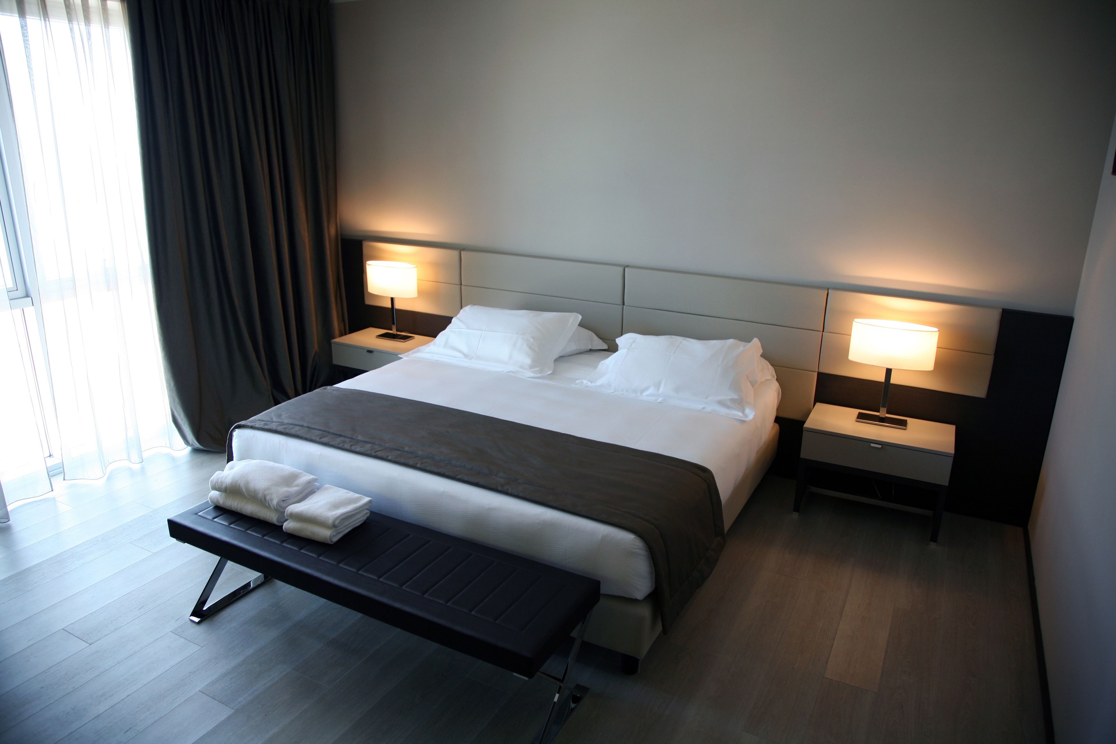 A modern hotel room with a large bed, two bedside tables with lamps, a bench, and a view of the outdoors.