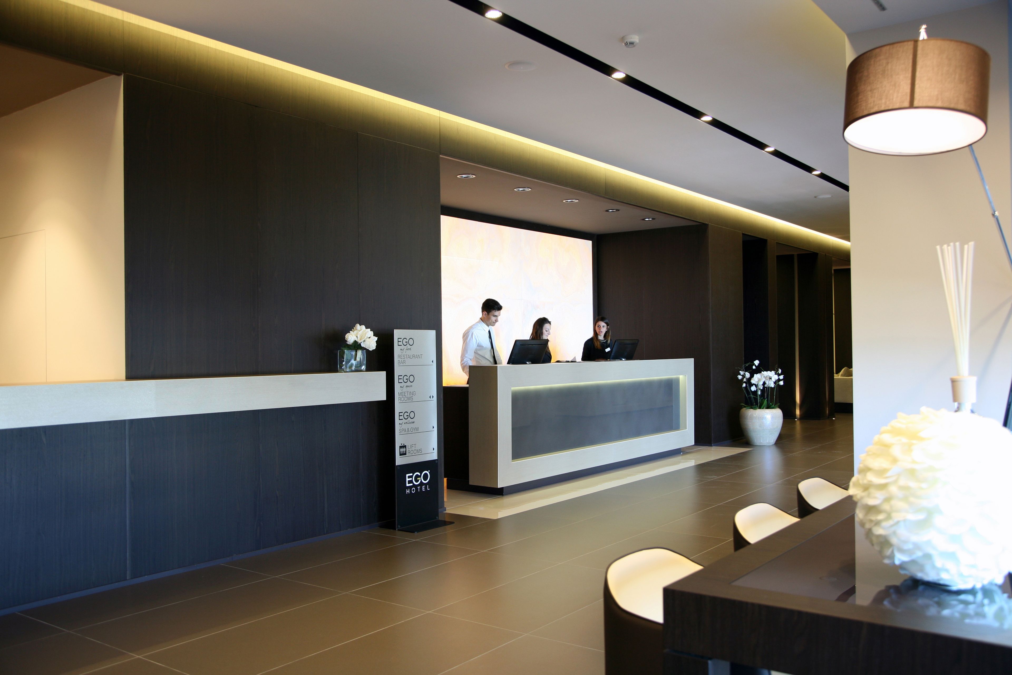 A modern hotel reception area with a reception desk, a digital screen, and a seating area with chairs.