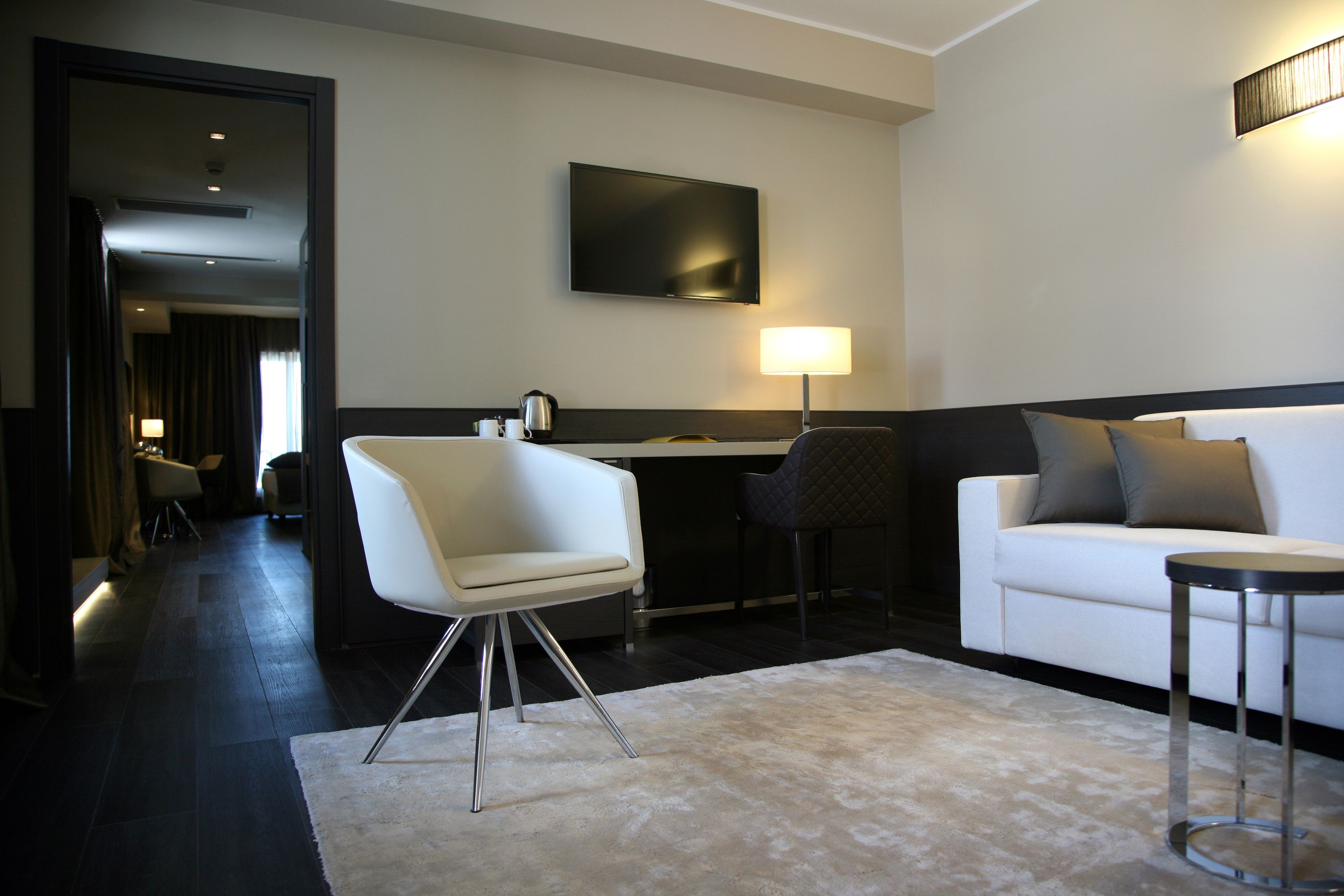 Ein modernes Hotelzimmer mit einer weißen Couch, einem kleinen runden Tisch, einem weißen Stuhl, einem an der Wand montierten Flachbildfernseher und einem Schreibtisch mit Lampe.