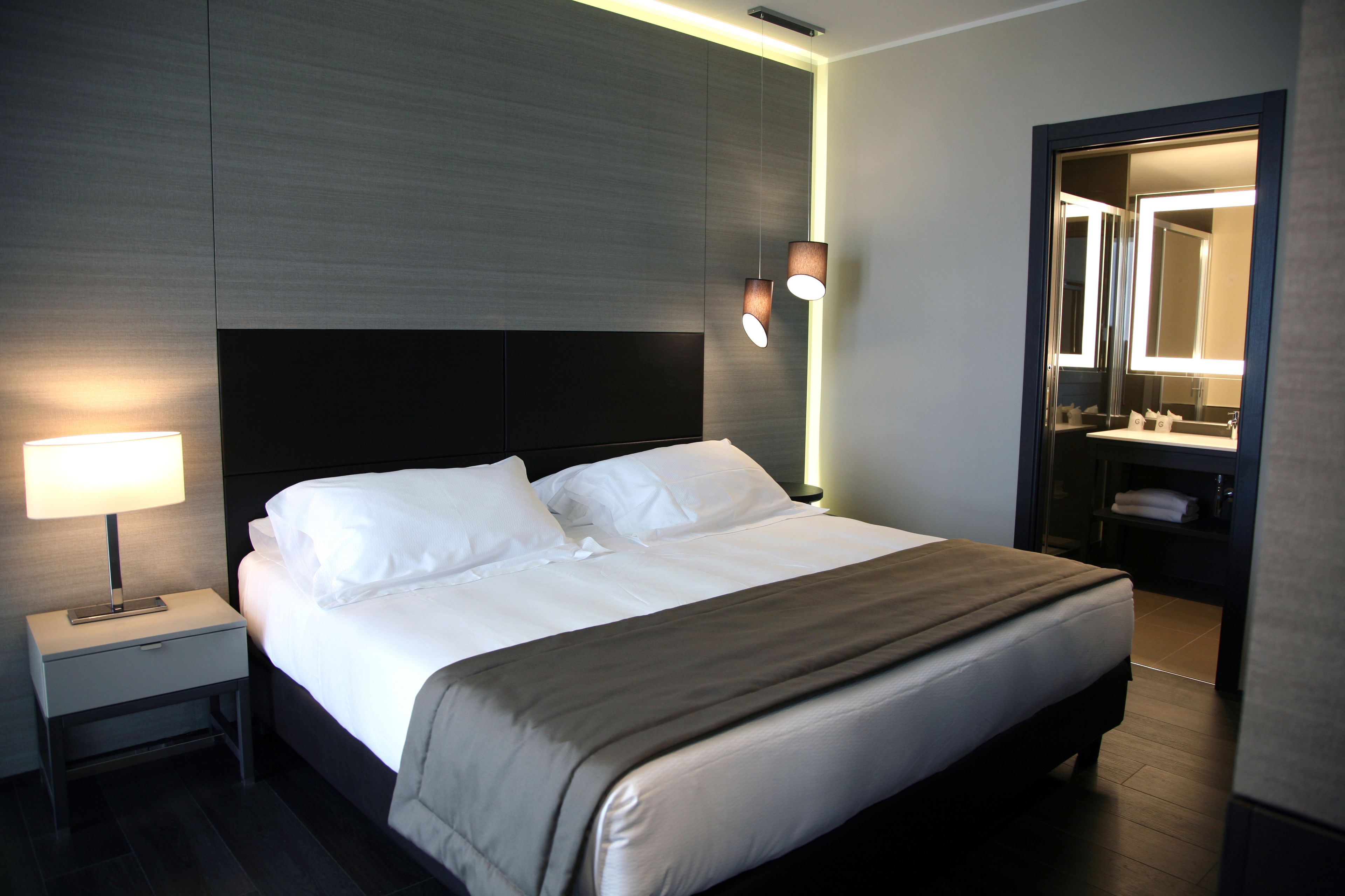 A modern hotel room with a large bed, bedside lamp, and a mirror above the sink.