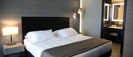 A modern hotel room with a large bed, bedside lamp, and a mirror above the sink.